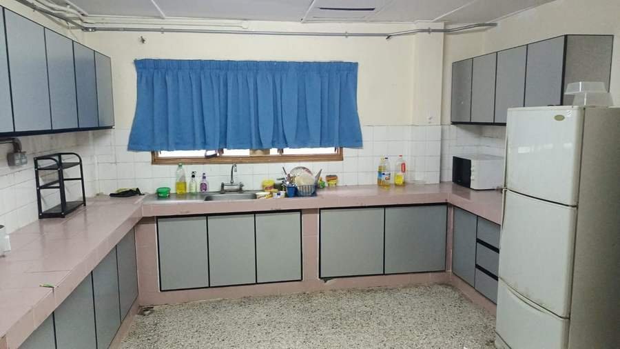 The kitchen in RKS 1. An L-shaped counter, a window with blue curtains, and a tall fridge at the end.