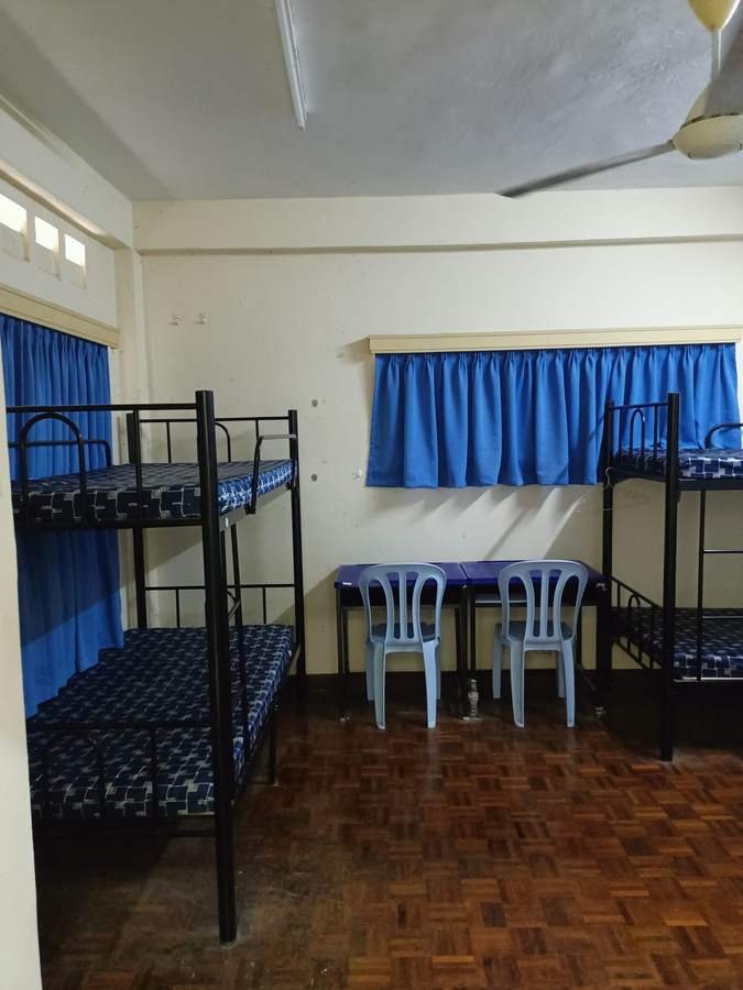 A bedroom in RKS 3. Two metal bunk beds with bare mattresses, blue curtains, a parquet floor and two plastic chairs.